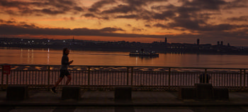 Mersey evening jogger This urban landscape photograph, taken in Liverpool, United Kingdom, captures a lone jogger running alongside the River Mersey during an autumn evening at dusk. The sky is filled with vibrant sunset hues, casting an orange glow over the water as lights begin to appear across the city skyline. Notable Liverpool landmarks are visible in the distance, including the towers of the iconic Liver Building, contributing to the unique character of the scene. A ferry crosses the River Mersey, illuminated as it travels, further emphasising the urban setting. The jogger along the waterfront is the main subject of the composition, embodying active city life against the backdrop of the tranquil river and dramatic sunset.